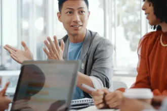 Asian man with black hair wearing a grey jacket with a powder blue shirt sitting in a conference room with his coworkers discussing how to grow their business with LegalZoom.