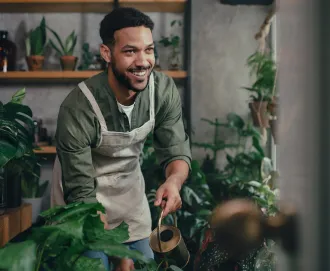 Black man with a short afro wearing a green shirt and brown apron smiling working in his greenhouse, watering plants after registering his trademark with LegalZoom.