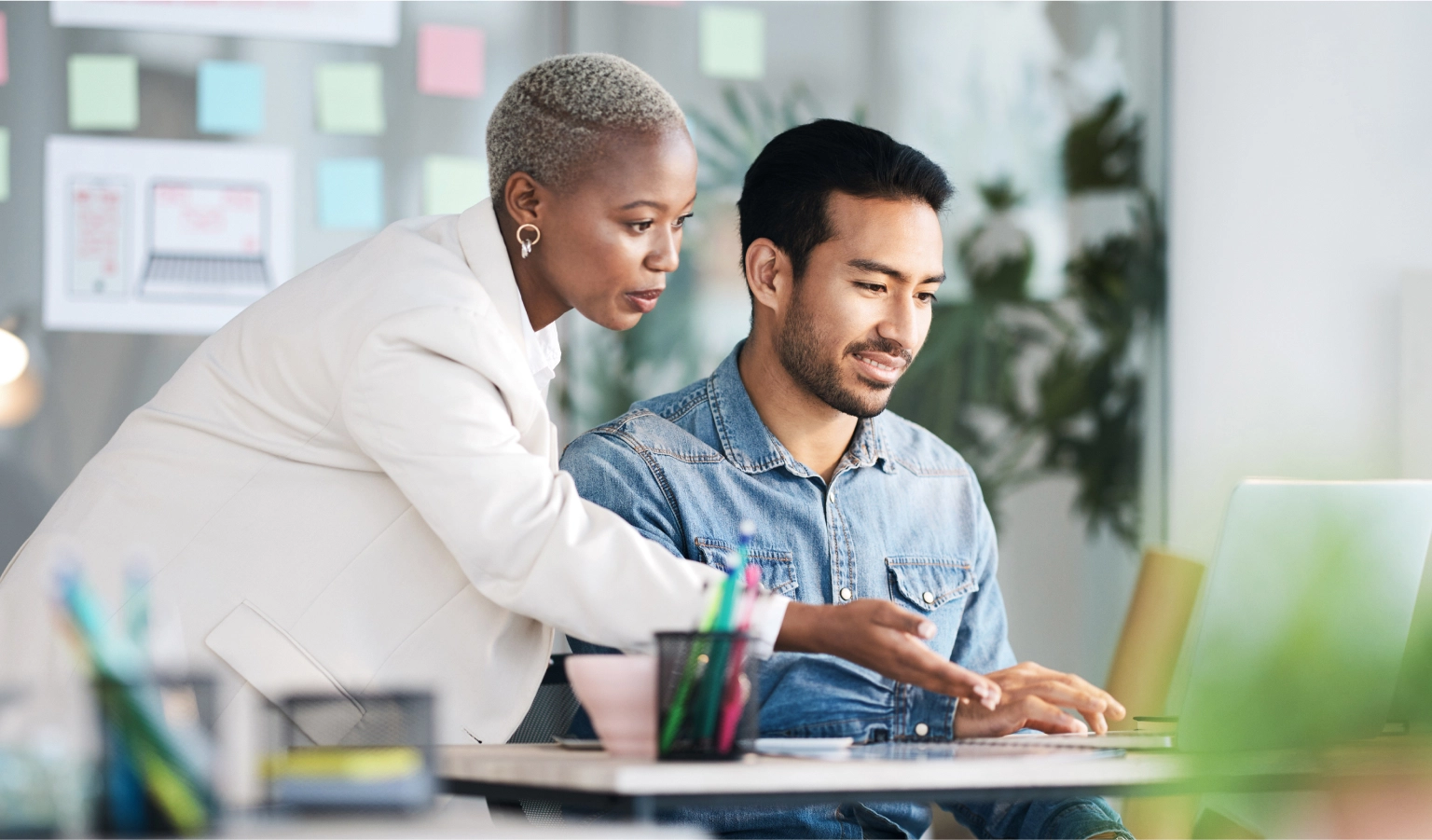 Black woman with short blonde afro wearing gold earrings and a white jacket leaning over the shoulder of an Asian man with black hair and short beard wearing a denim shirt looking at ways they can grow their business with LegalZoom partners on a laptop