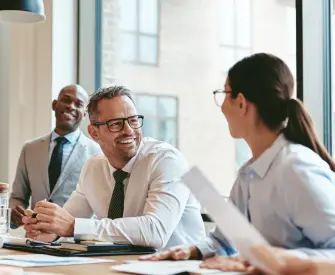 A man wearing glasses and a white button-up shirt and tie is smiling at a brunette woman wearing glasses in a conference room. In the background is smiling as they discuss how to incorporate their business.