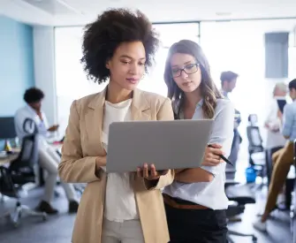 Black woman with an afro wearing a tan business jacket and white shirt holding her laptop, talking to a female colleague with dark hair wearing glasses and a white button up shirt discussing how to incorporate their business with LegalZoom.