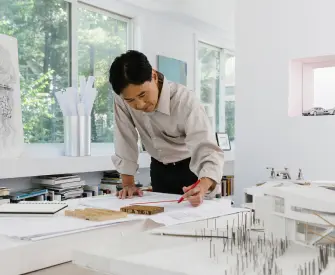 Smiling man in an off-white shirt and black pants leaning over a drafting table in a sunlit office as he works on blueprints for his newly formed LLC.