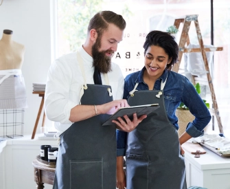 Woman with dark hair wearing a denim shirt and a grey apron looking at a LegalZoom LLC on a tablet with her business partner, a man with a beard wearing a white shirt and grey apron.